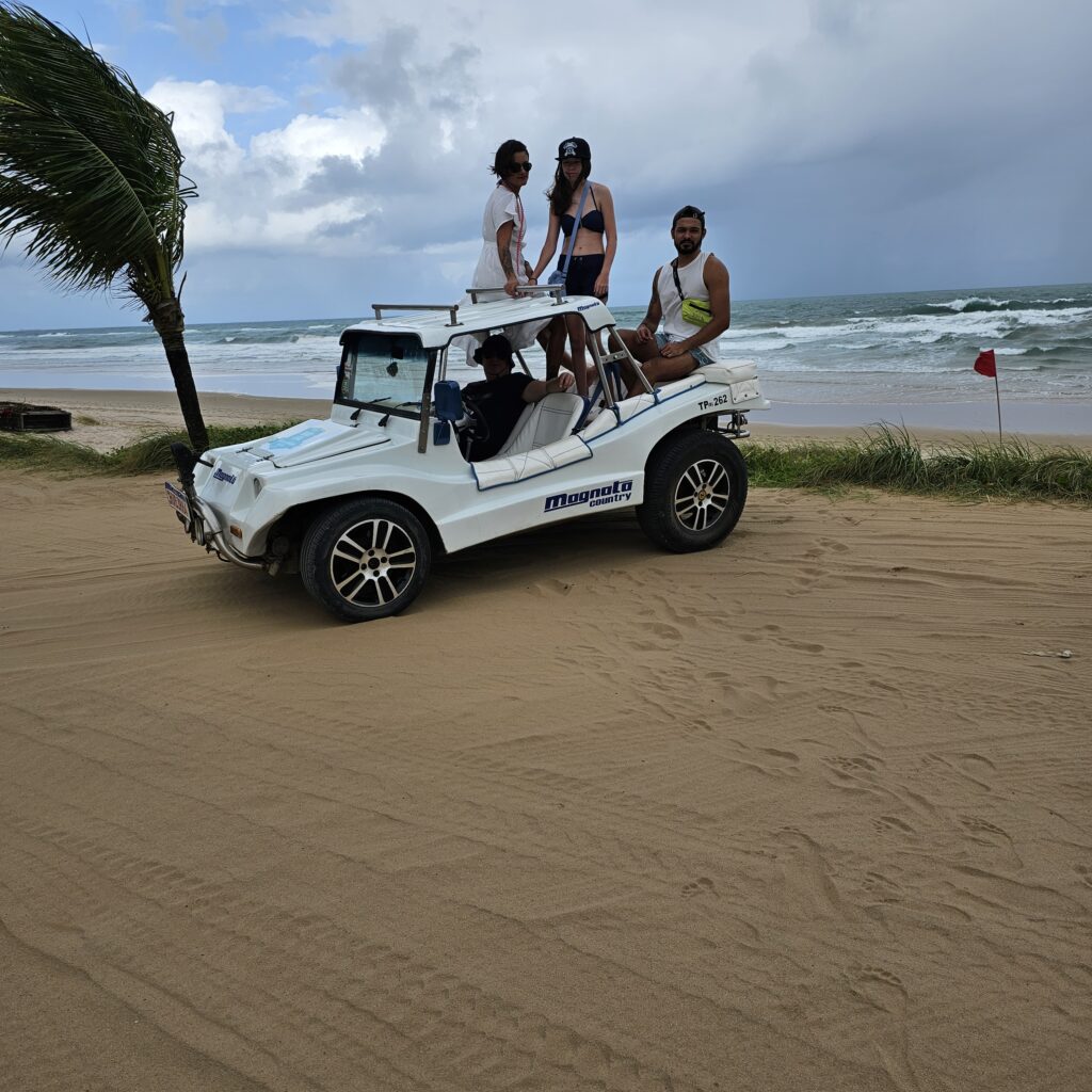 Vier Personen sitzen und stehen auf einem weißen Strandbuggy am Strand, im Hintergrund Palme und aufgewühltes Meer