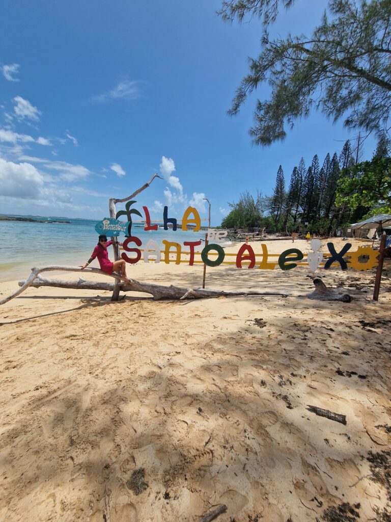 Frau sitzt auf einem Treibholzast vor dem bunten Schriftzug "Ilha de Santo Aleixo" am Strand