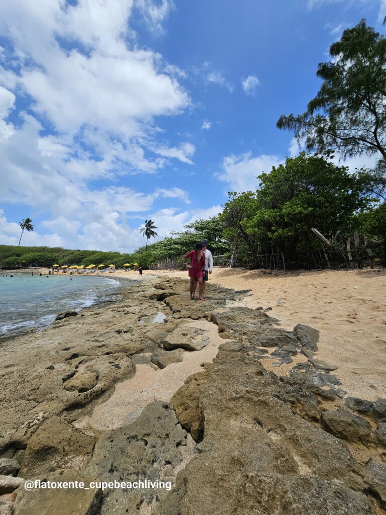 Paar steht auf Felsen am Strand der Ilha de Santo Aleixo, im Hintergrund Meer und gelbe Sonnenschirme