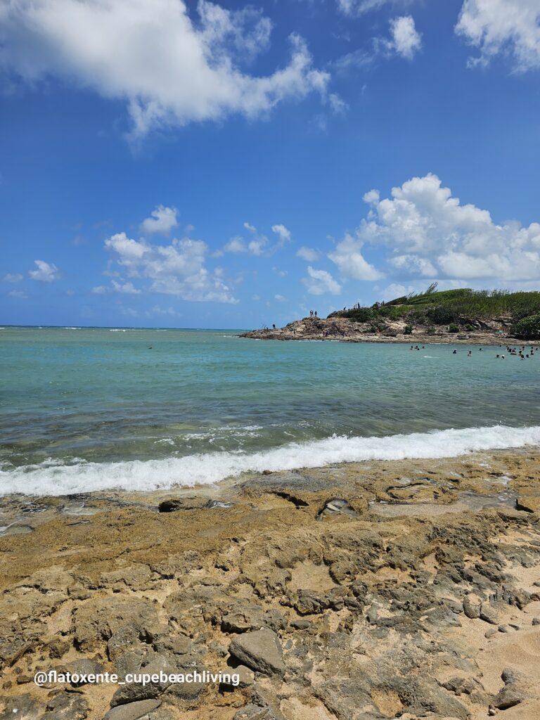 Türkisfarbenes Meer mit Wellen an einer Felsküste, im Hintergrund Menschen auf Felsen und im Wasser bei der Ilha de Santo Aleixo