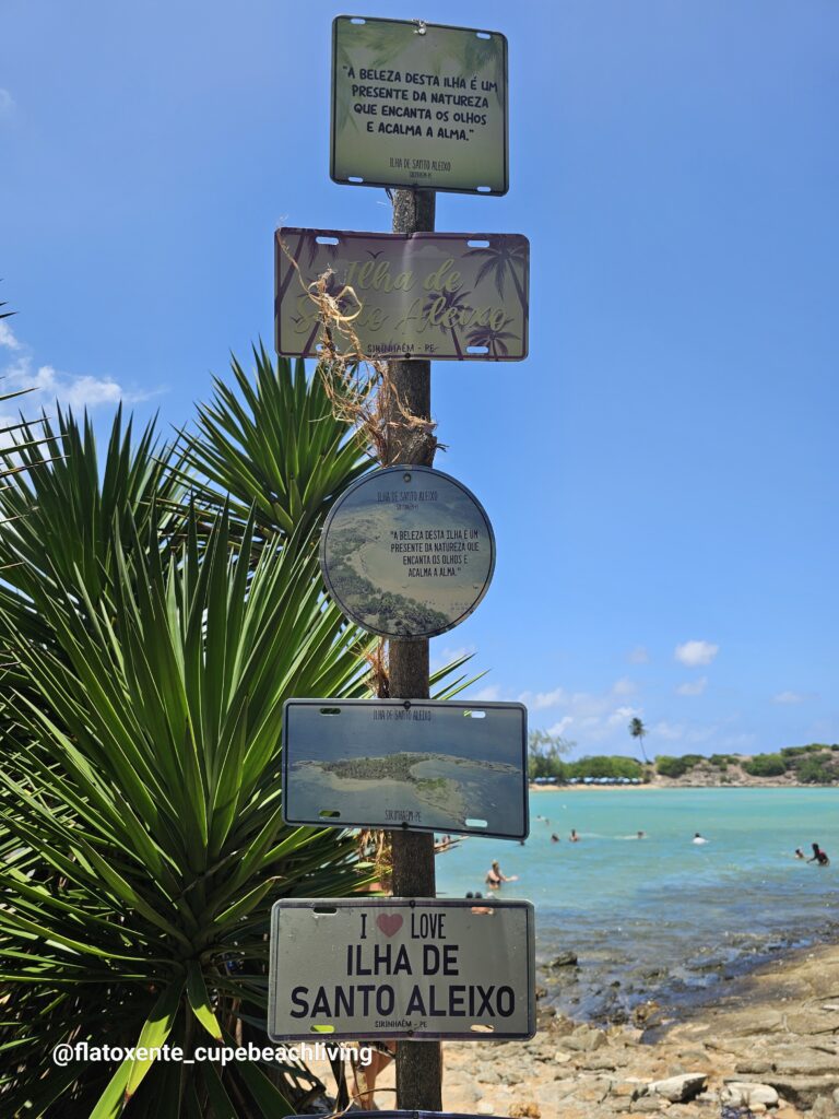 Mehrere Nummernschild-artige Schilder mit der Aufschrift "Ilha de Santo Aleixo" an einem Pfosten, im Hintergrund Strand und Meer