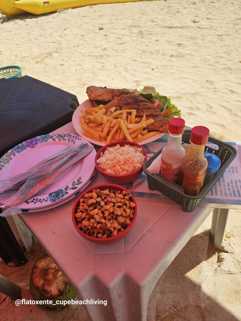 Teller mit gegrilltem Fisch, Pommes und Salat neben Schälchen mit Reis und Bohnen auf einem Tisch am Strand