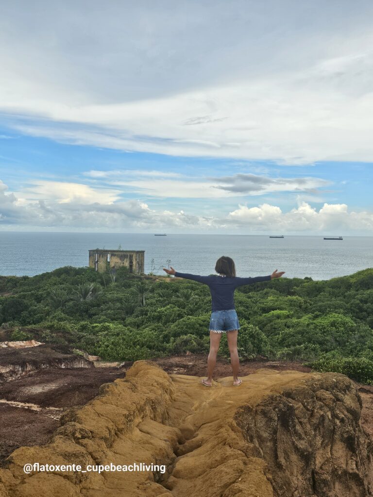 Frau mit ausgebreiteten Armen auf einem Felsen am Mirante Vila Velha, im Hintergrund eine alte Ruine und Schiffe auf dem Meer