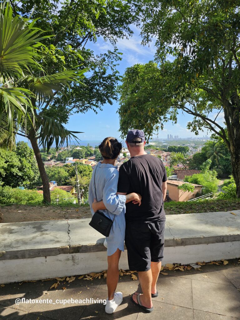 Paar von hinten umarmt sich an einem Aussichtspunkt in Olinda unter Bäumen, Blick auf Dächer, Meer und die Skyline von Recife