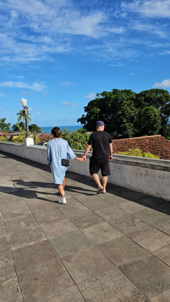 Paar geht Hand in Hand auf einer Terrasse in Olinda entlang, im Hintergrund Kolonialdächer und das Meer