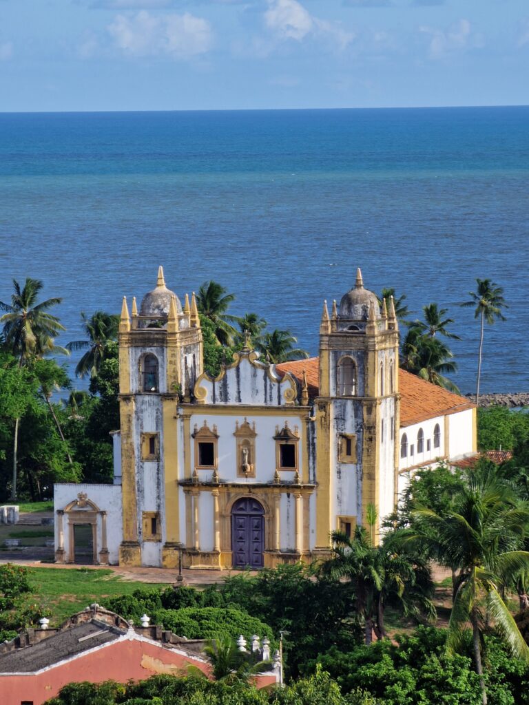 Weiß-gelbe Kolonialkirche mit zwei Türmen direkt am Meer in Olinda, umgeben von Palmen