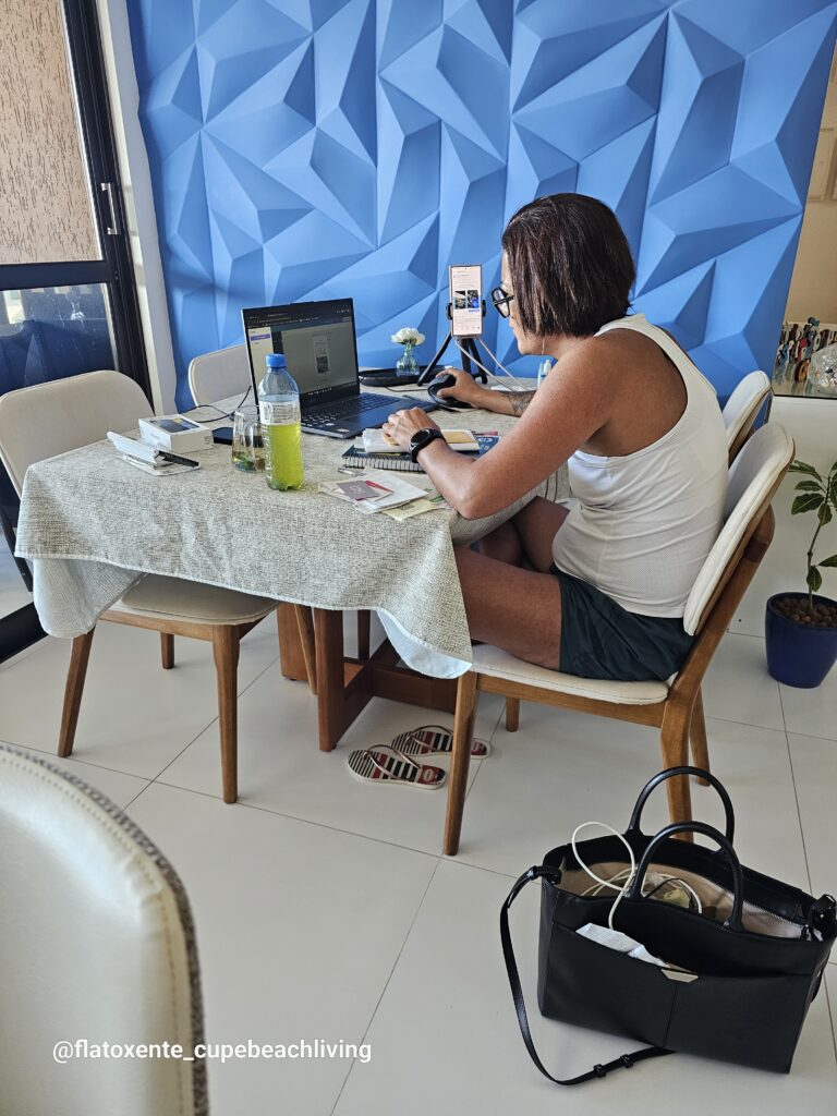 Frau sitzt am Esstisch und arbeitet am Laptop, daneben ein Smartphone auf einem Stativ, im Hintergrund eine blaue 3D-Wandverkleidung