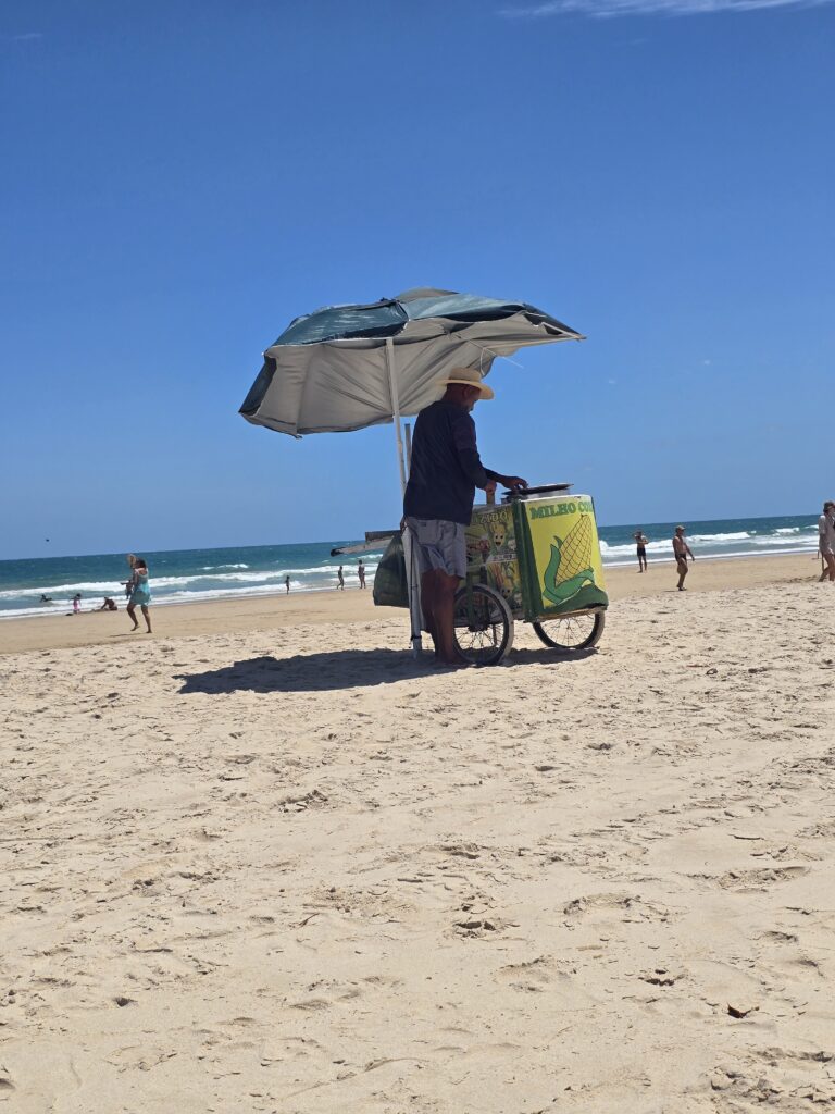 Maisverkäufer mit Sonnenschirm und Verkaufswagen "Milho Cozido" am Sandstrand von Praia do Cupe