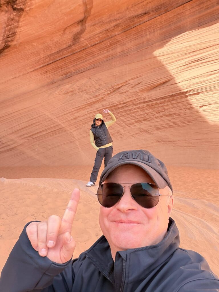 Selfie eines Paares in Sonnenbrillen und Mützen vor der roten Sandsteinwand von Bidii the Arch