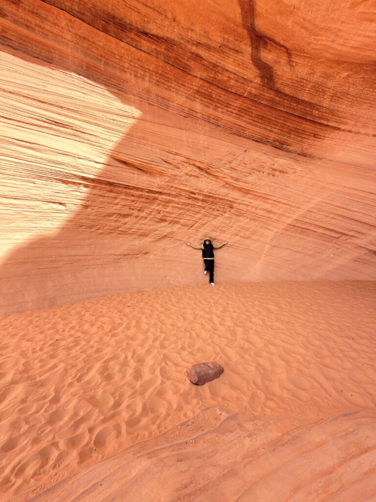Person posiert an geschwungener roter Sandsteinwand über einer Sanddüne bei Bidii the Arch
