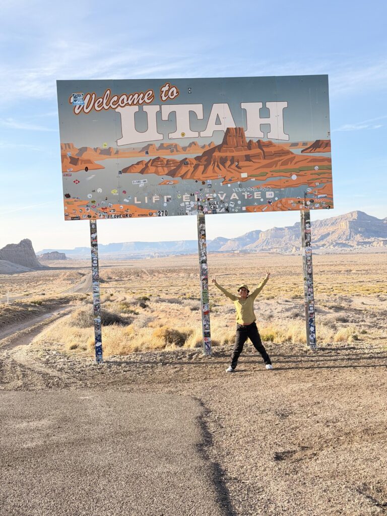 Person posiert jubelnd vor dem "Welcome to Utah – Life Elevated" Schild in der Wüstenlandschaft