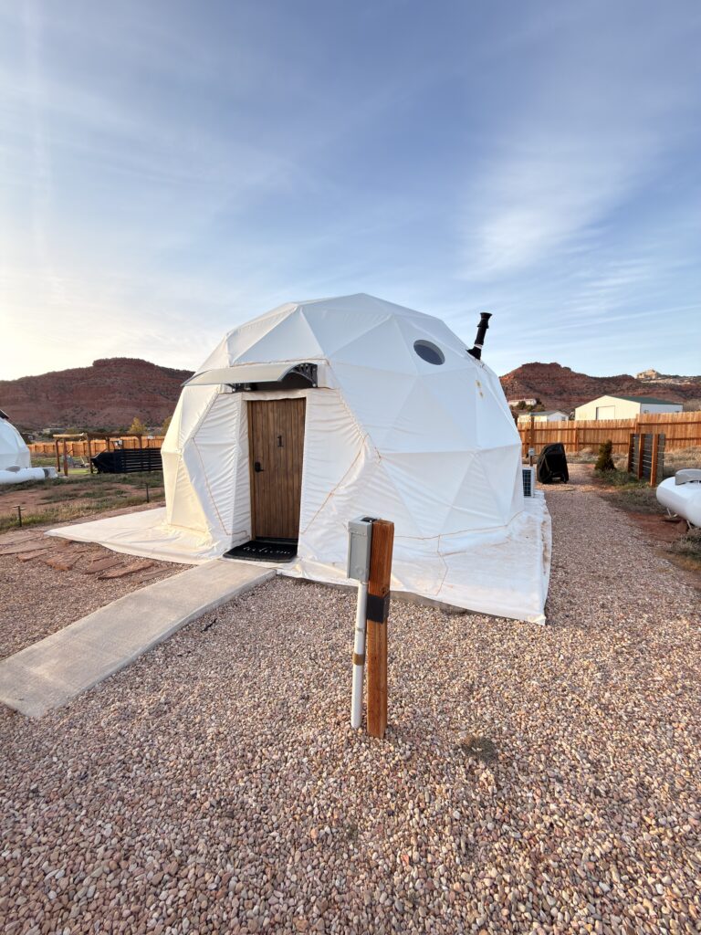 Weißes Glamping-Kuppelzelt White Camel in Kanab, Utah mit Holztür und rotem Felsen im Hintergrund