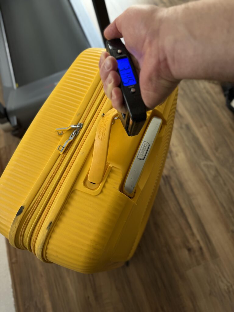 Hand hält eine digitale Kofferwaage mit blauem Display am Griff eines gelben American Tourister Koffers