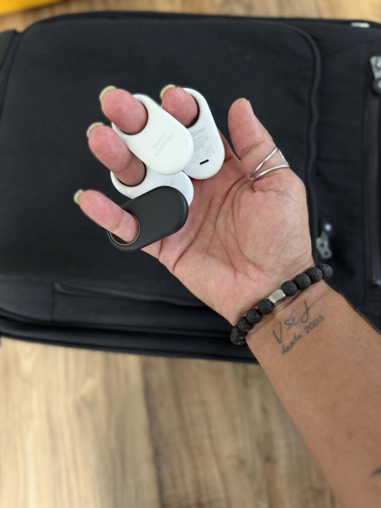 Hand hält vier Samsung Galaxy SmartTag2 Tracker in Weiß und Schwarz vor schwarzem Rucksack auf Holzboden