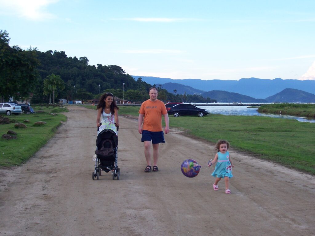Familie spaziert bei Abendlicht auf einem unbefestigten Weg am Wasser in Paraty, im Hintergrund Berge, Brasilien