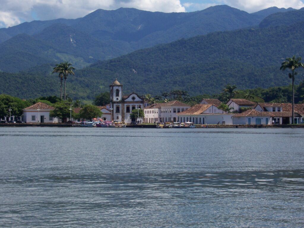 Blick vom Wasser auf die weiße Altstadt von Paraty mit Kirchturm vor bewaldeten Bergen, Brasilien