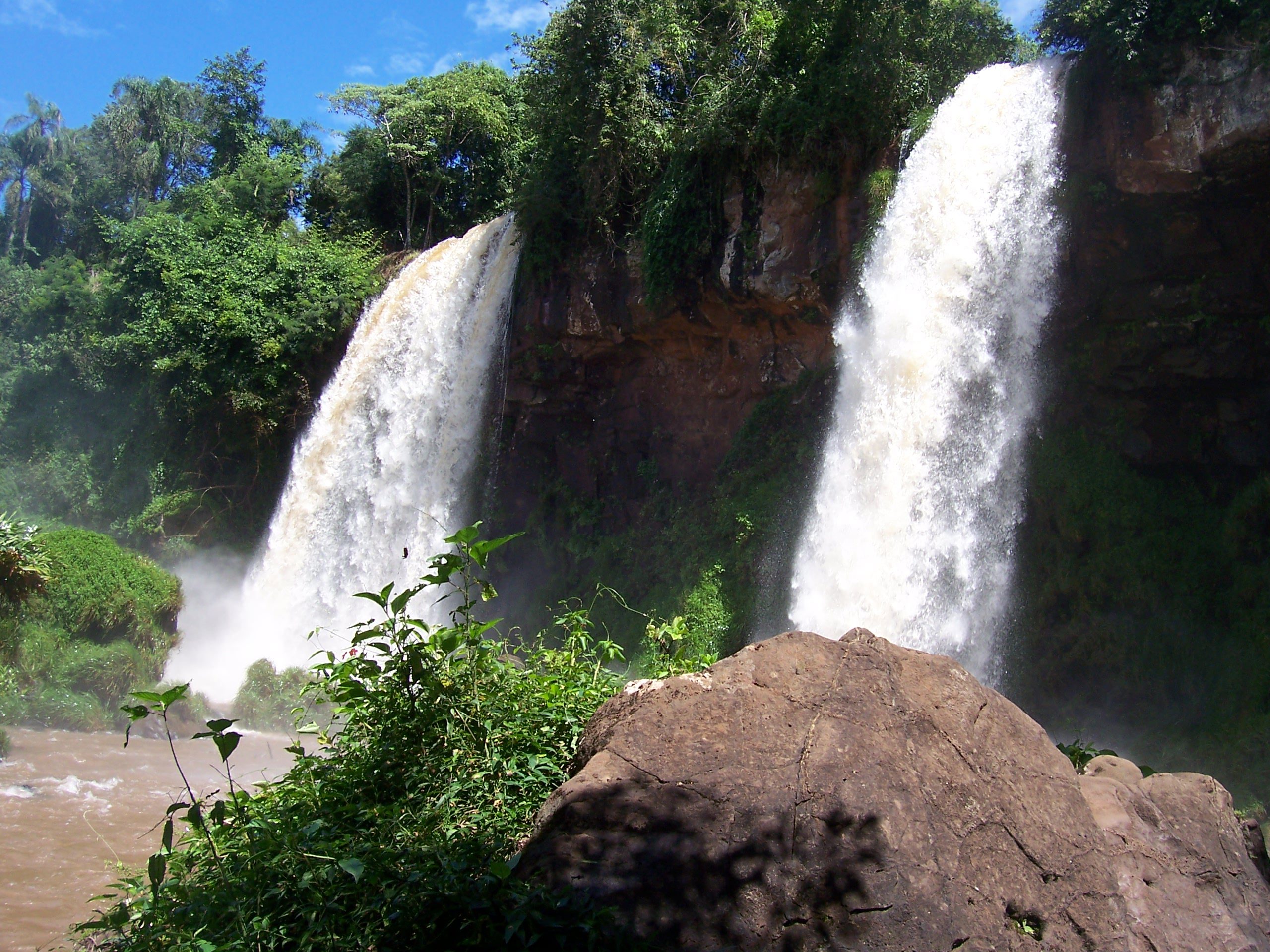 Nahaufnahme zweier nebeneinander stürzender Wasserfälle mit Felsen im Vordergrund, Iguazú-Fälle, Argentinien
