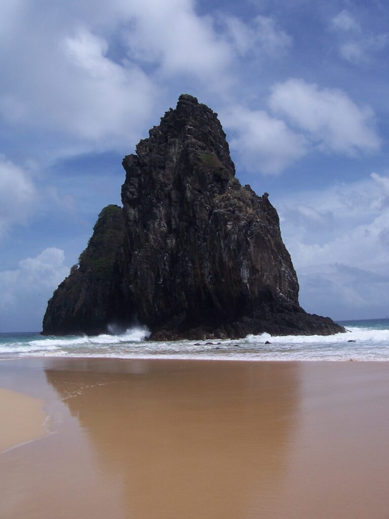 Steil aufragender schwarzer Felsturm im türkisfarbenen Meer, vom Strand aus fotografiert, Fernando de Noronha, Brasilien