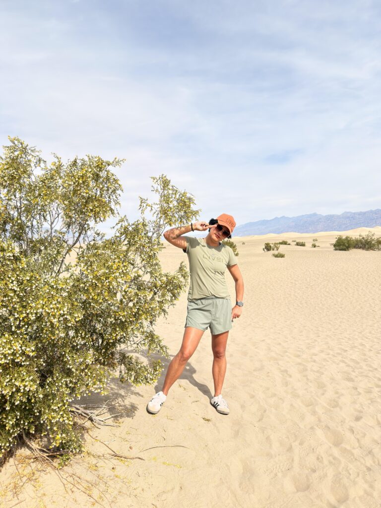 Frau in Wüstenoutfit posiert an einem blühenden Wüstenstrauch vor den Mesquite Flat Sand Dunes