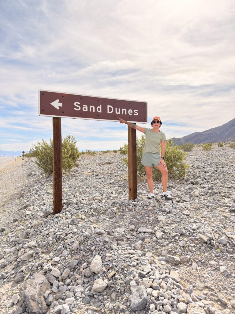 Frau steht neben einem Schild "Sand Dunes" auf steinigem Untergrund im Death Valley