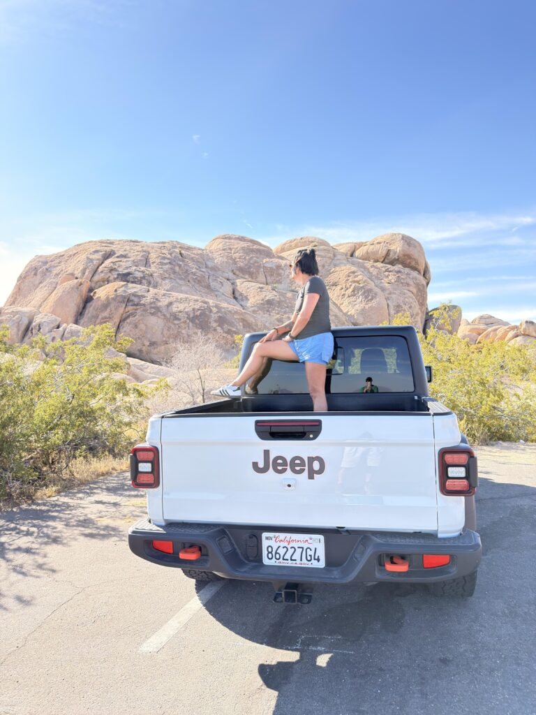 Frau sitzt auf der Ladefläche eines Jeeps vor Felsformationen im Joshua Tree National Park