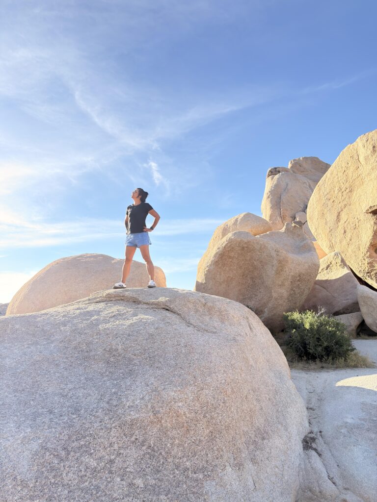 Frau steht auf einem Felsblock im Joshua Tree National Park und blickt in den Himmel
