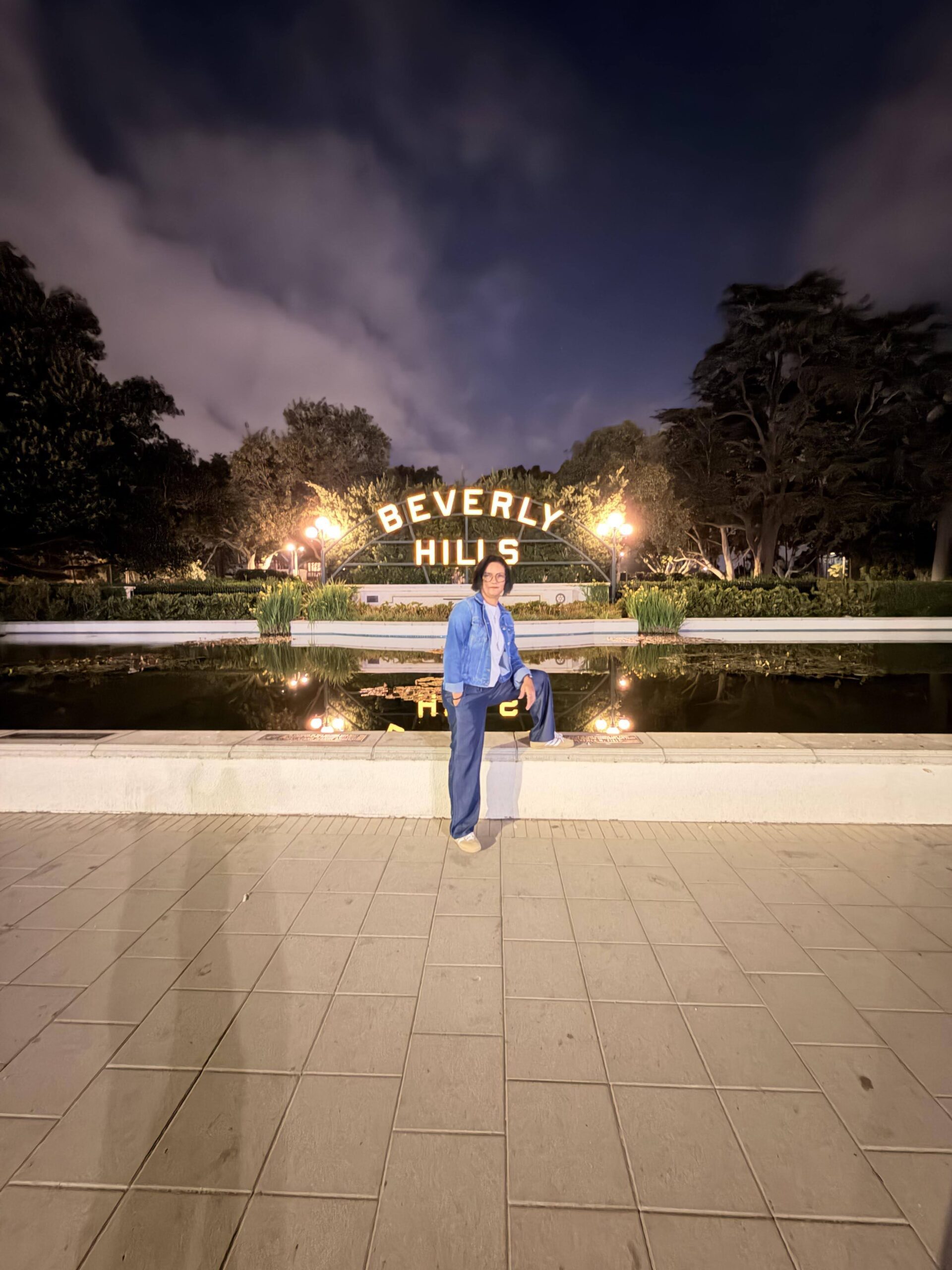 Frau sitzt nachts auf einer Mauer vor dem beleuchteten Beverly Hills Schild mit Wasserspiegelung