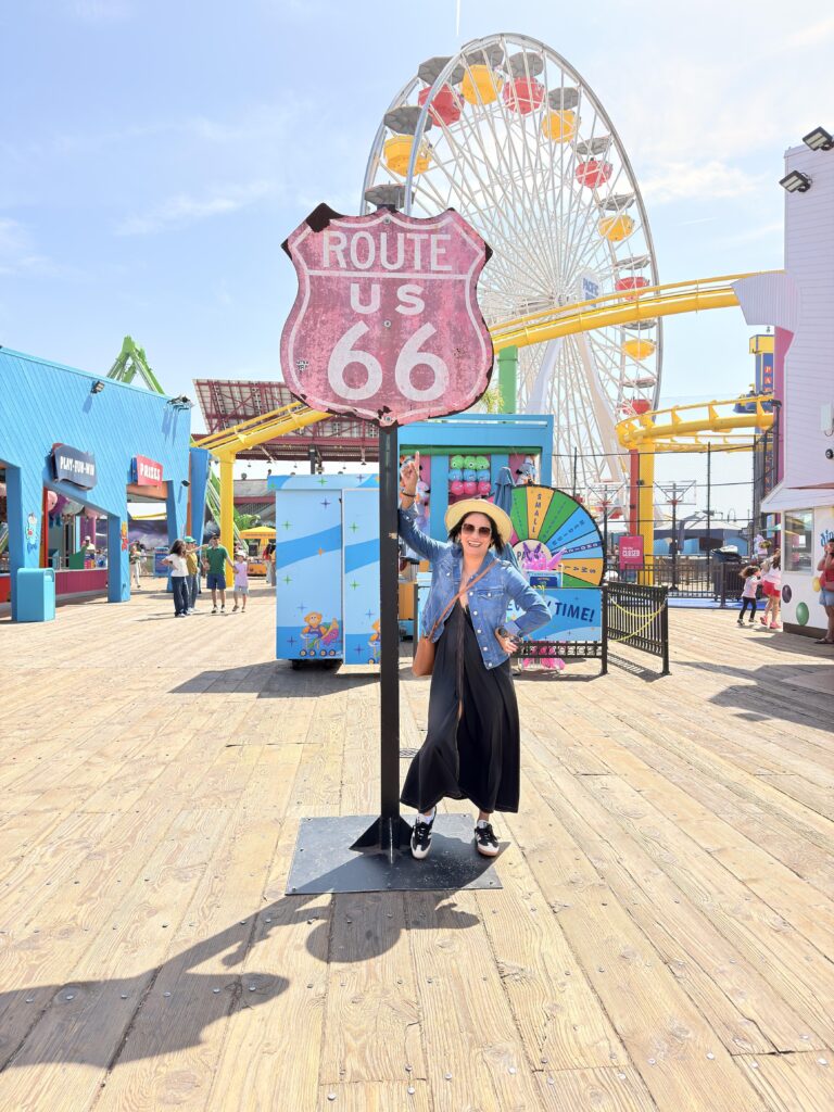 Frau steht lächelnd unter einem Route-US-66-Schild vor einem bunten Riesenrad im Pacific Park