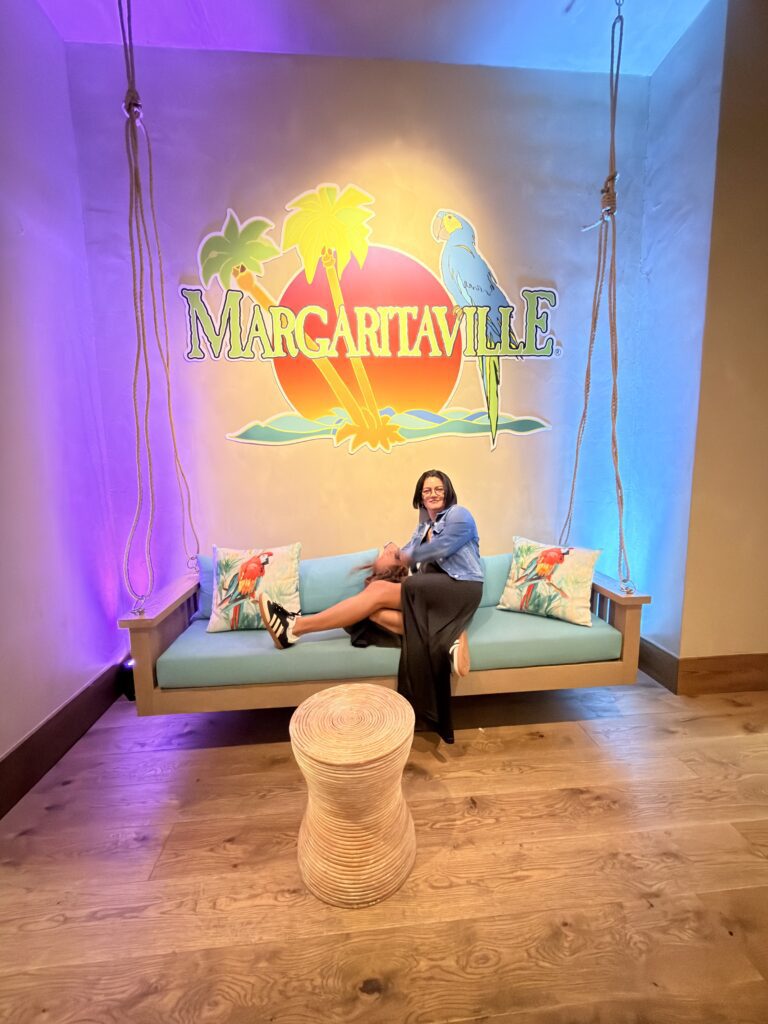 Frau sitzt auf einer Hängeschaukel vor dem bunten Margaritaville-Logo mit Papagei und Palmen