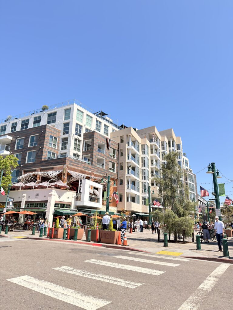 Moderne Wohn- und Geschäftsgebäude in Downtown San Diego bei blauem Himmel, Zebrastreifen im Vordergrund, Passanten und Straßencafés mit amerikanischen Flaggen